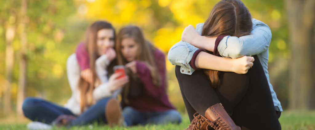 A girl sits alone with her head down while two girls in the background look at a phone together in an outdoor setting.