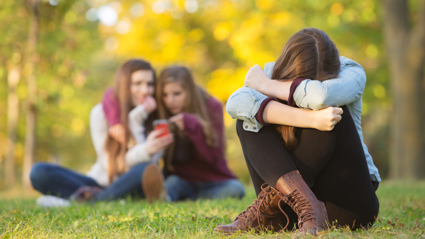 A girl sits alone with her head down while two girls in the background look at a phone together in an outdoor setting.